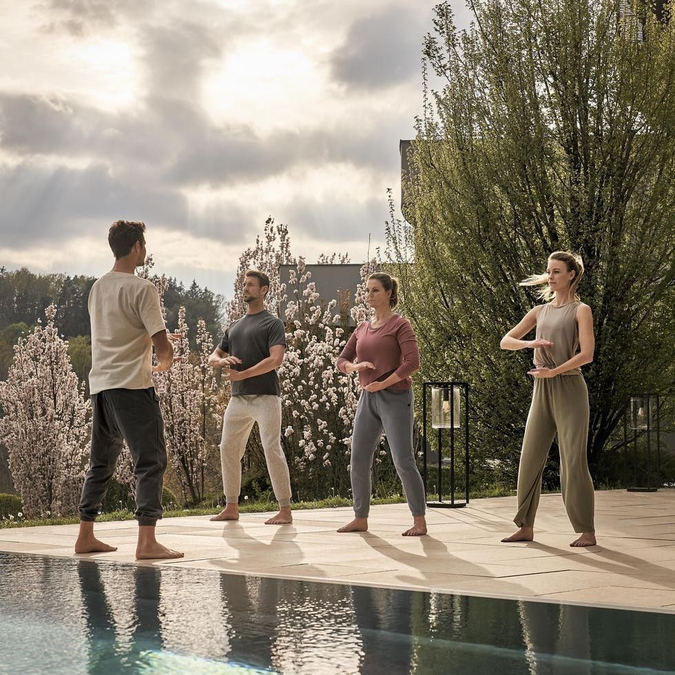 Four people practicing yoga by a pool with trees and a house in the background.