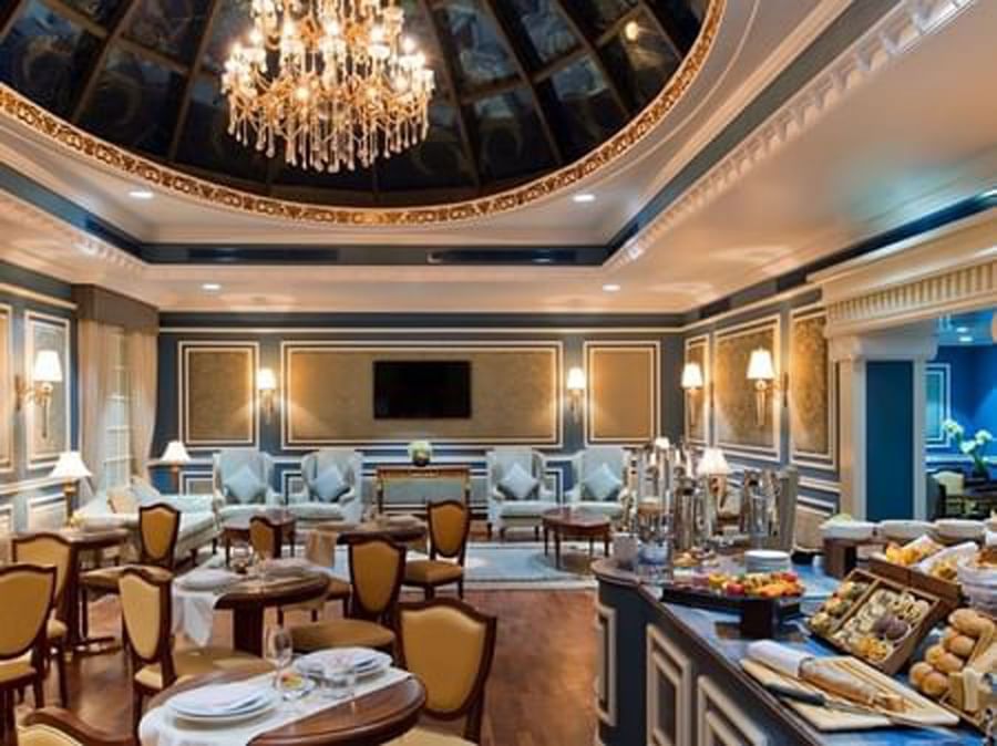 Elegant lounge featuring tan chairs by a breakfast buffet under a crystal chandelier in Coral Room at Warwick Doha Hotel
