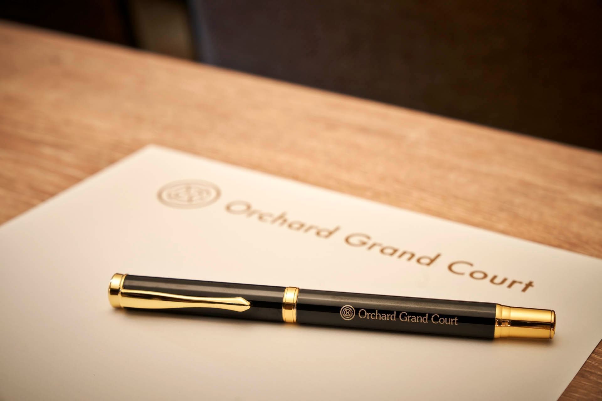 A black pen with gold accents rests on a beige notepad featuring the Orchard Grand Court logo