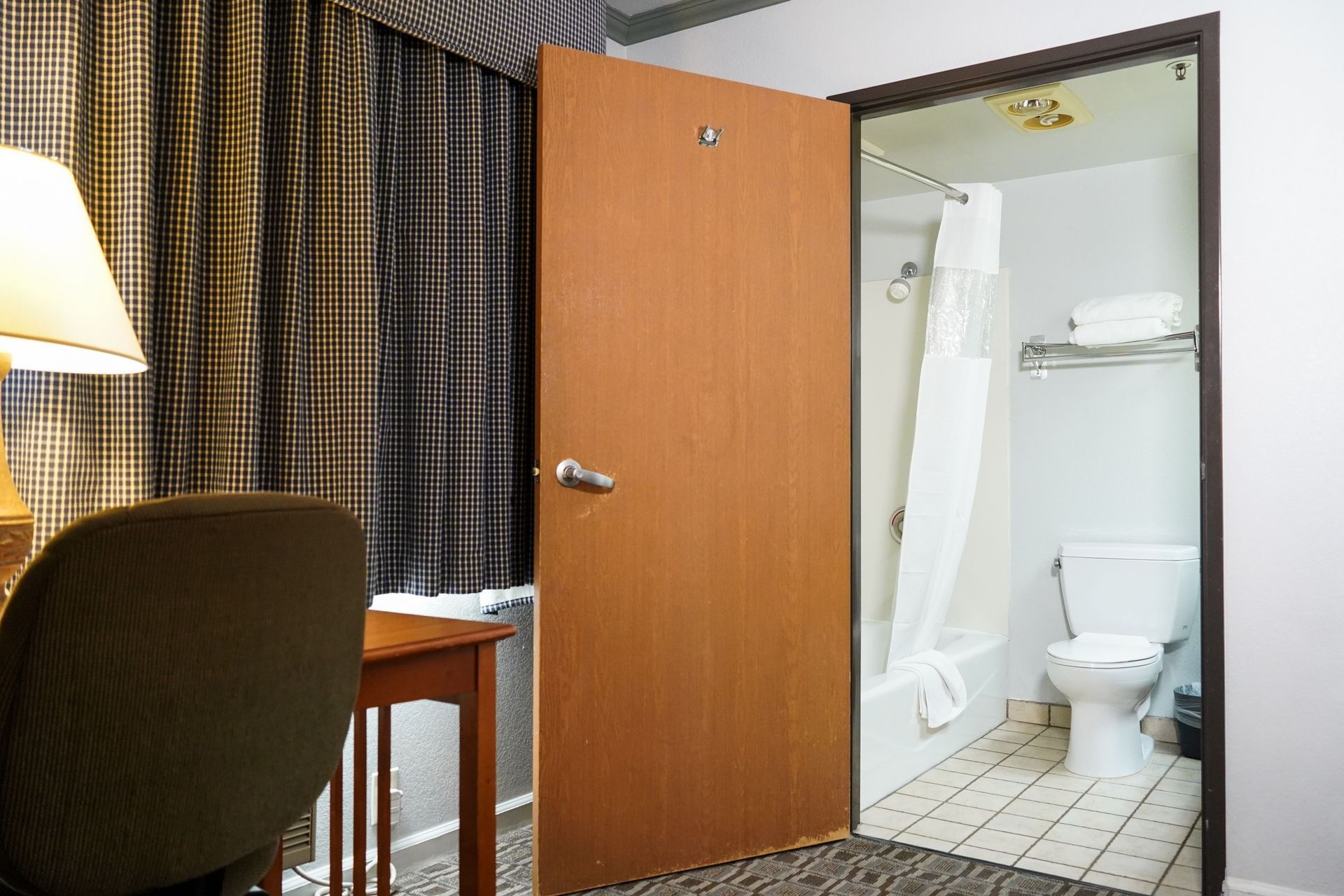 A hotel room with a desk, chair, and lamp, with an open door to a bathroom with a toilet, shower, and bathtub.