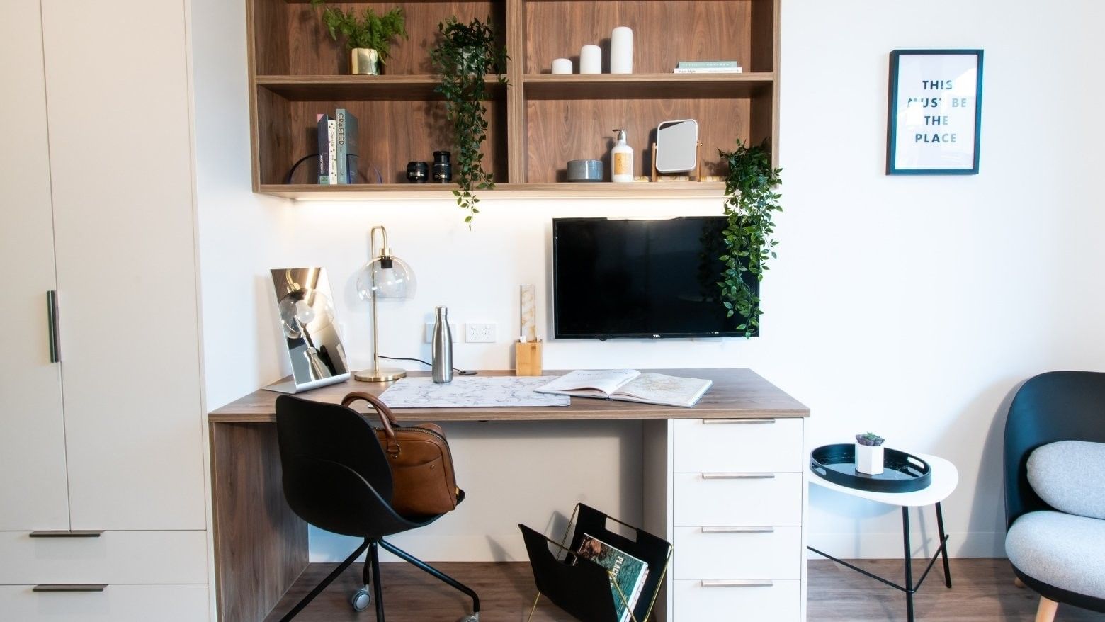 Office setup with desk, chair, computer monitor, framed wall art, and shelves with plants and decor.