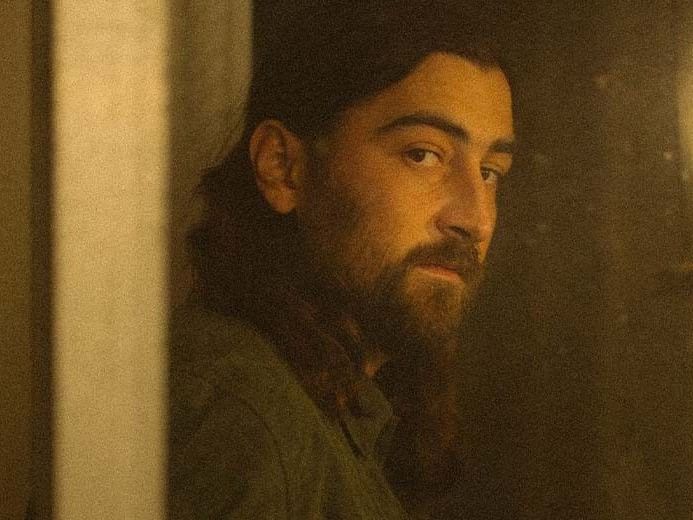 A bearded man with long hair looks pensively out of a doorway at Noah Kahan’s concert.