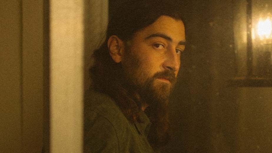 A bearded man with long hair looks pensively out of a doorway at Noah Kahan’s concert.