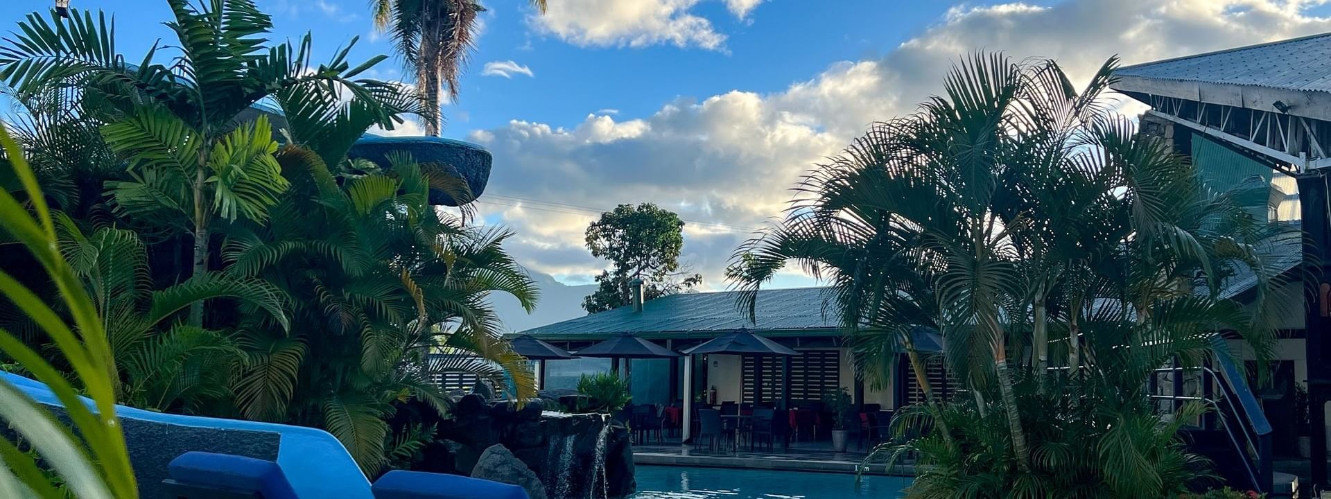 Blue lounge chairs by a swimming pool under fluffy white clouds surrounding palm trees at Tokatoka Resort in Nadi, Fiji