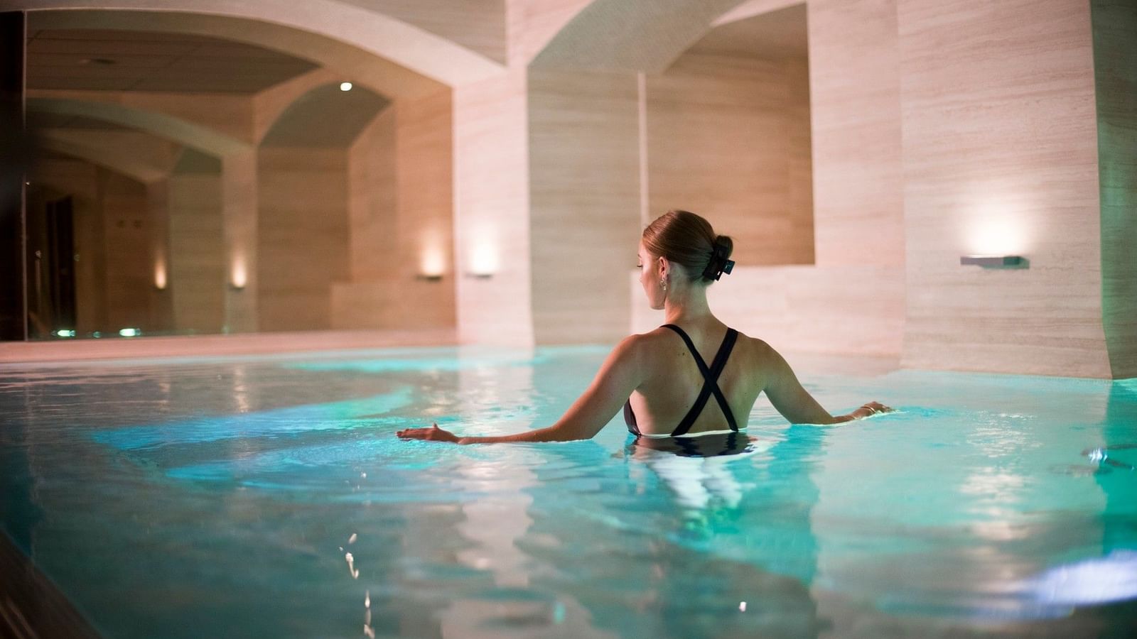 Kvinna kopplar av i upplyst inomhuspool med elegant arkitektur och mjuk belysning i spa-miljö
