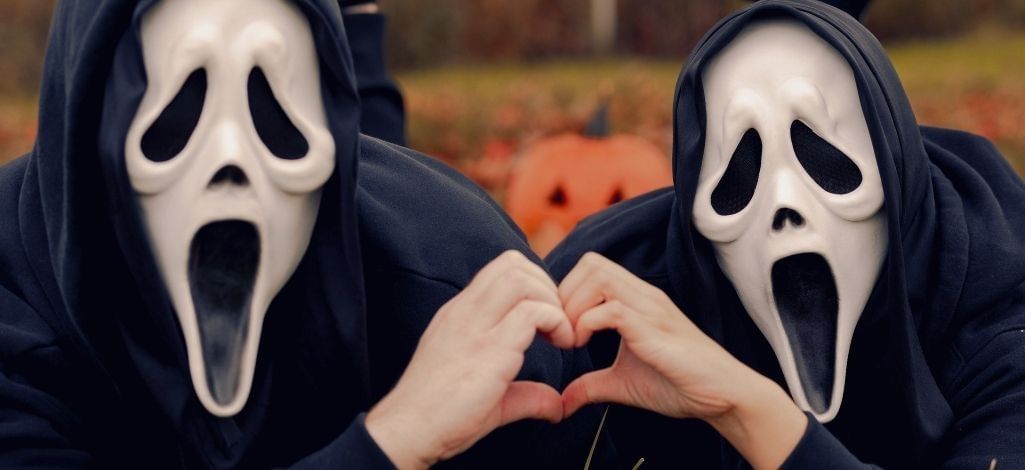 Two people in Ghostface costumes form a heart shape with their hands outdoors.