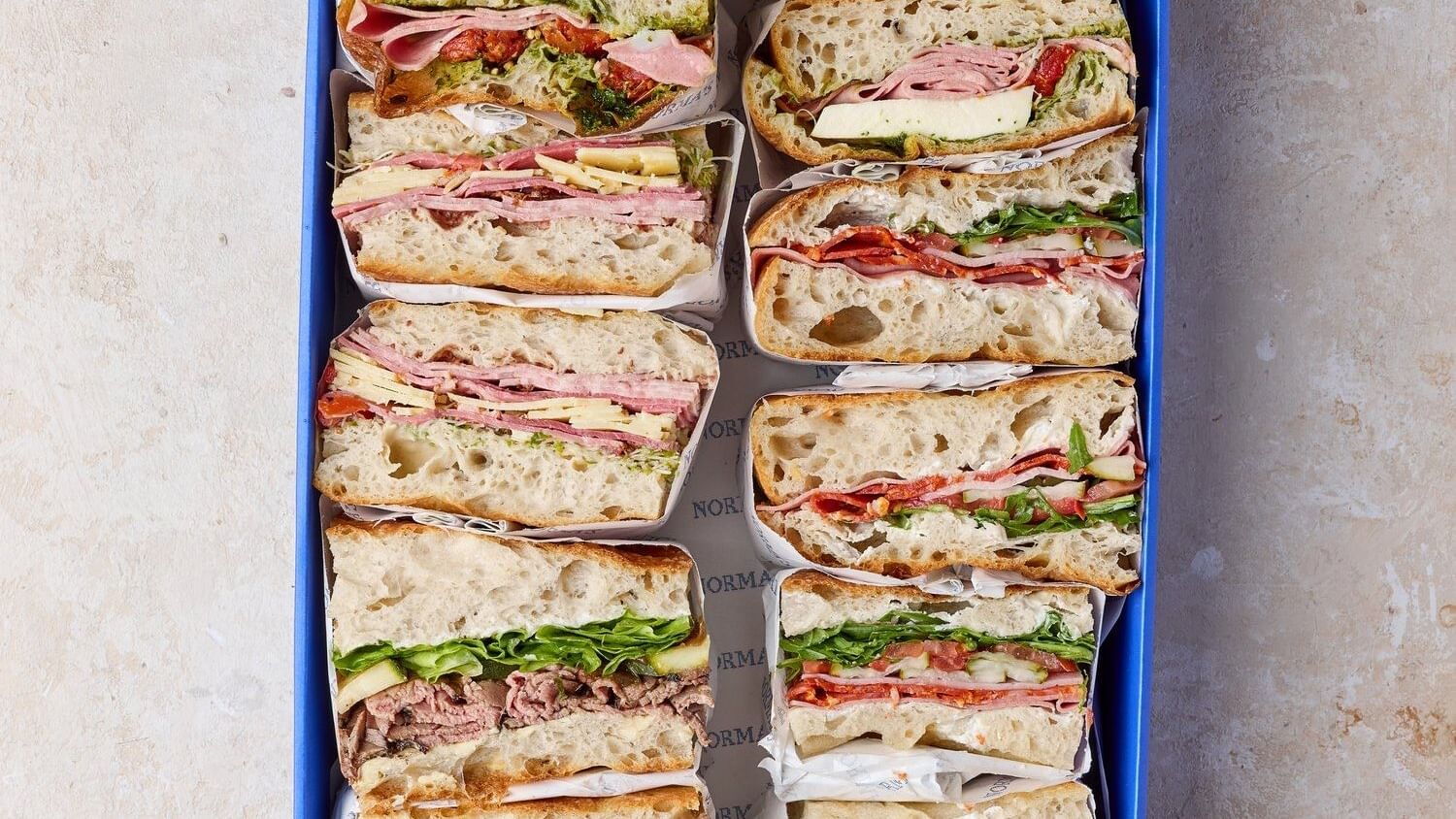 Assorted gourmet sandwiches in a blue box under bright light, surrounding fresh wraps in Norma\’s Deli at Elysium Manly