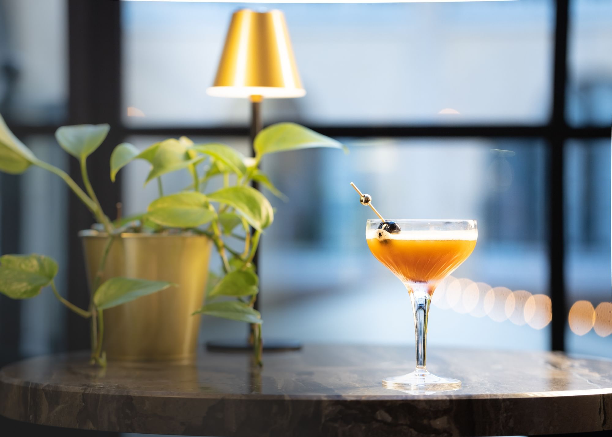 Cherry-garnished cocktail on a round table beside a potted plant and golden lamp at El Prado Hotel