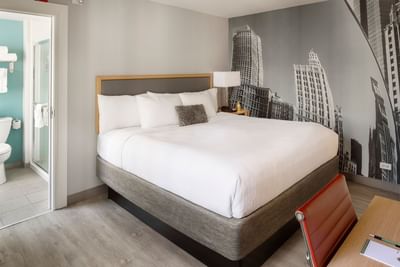 White king bed beside a nightstand and lamp under a Chicago skyline mural in King Suite at Hotel Saint Clair