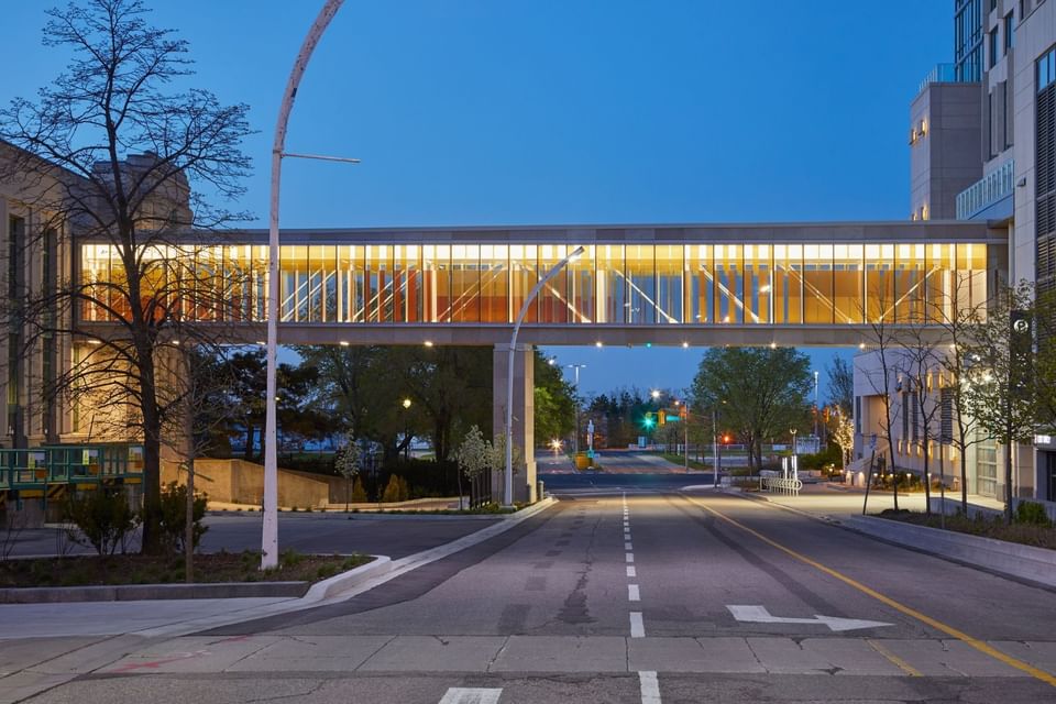 Illuminated glass skybridge connecting buildings above a quiet street, under a dusk sky at Hotel X Toronto