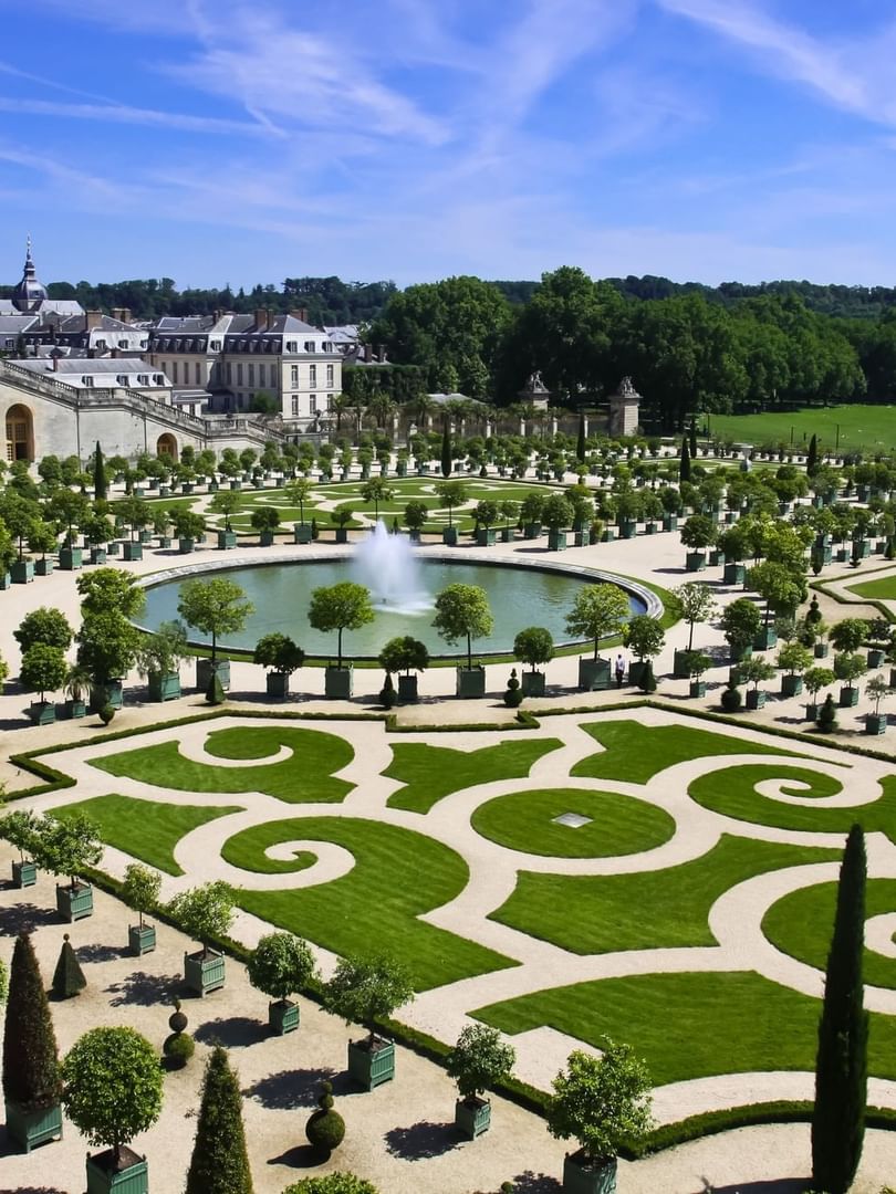 Round fountain by manicured lawns under a blue sky near the palace facade at Warwick Paris Champs Elysées