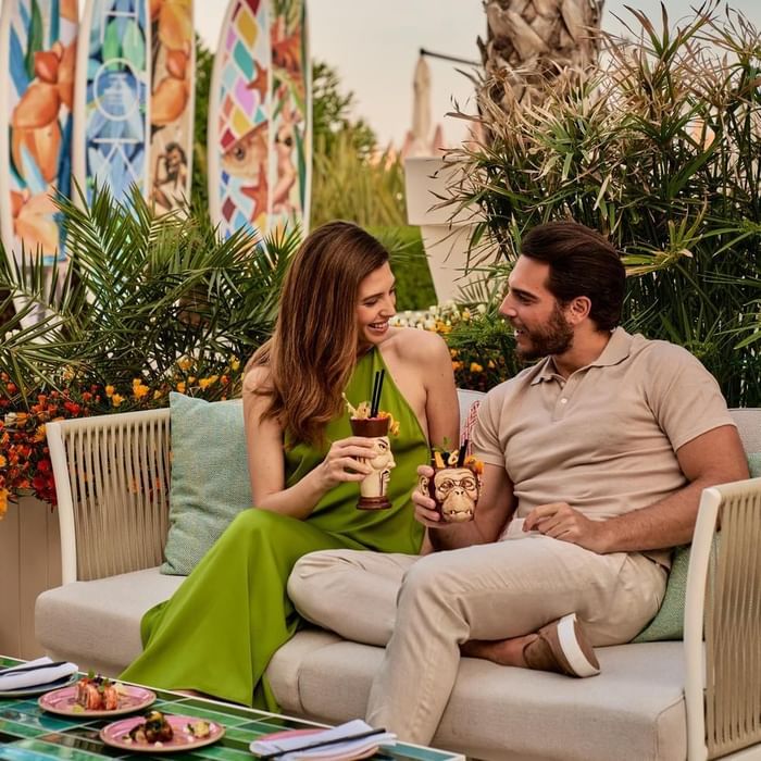 Coppia seduta su divano esterno, con drink esotici e cibo, circondati da piante e decorazioni tropicali.