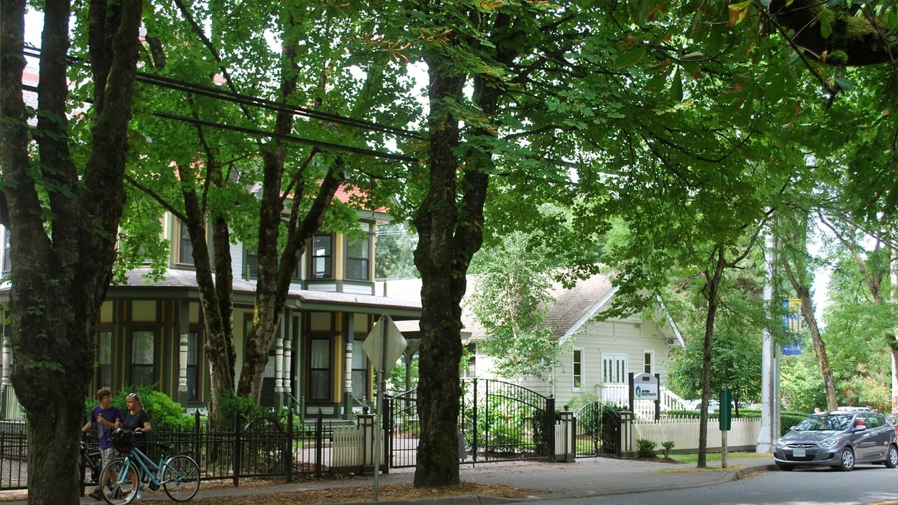Two people walk bikes near fenced homes with trees and a parked car.