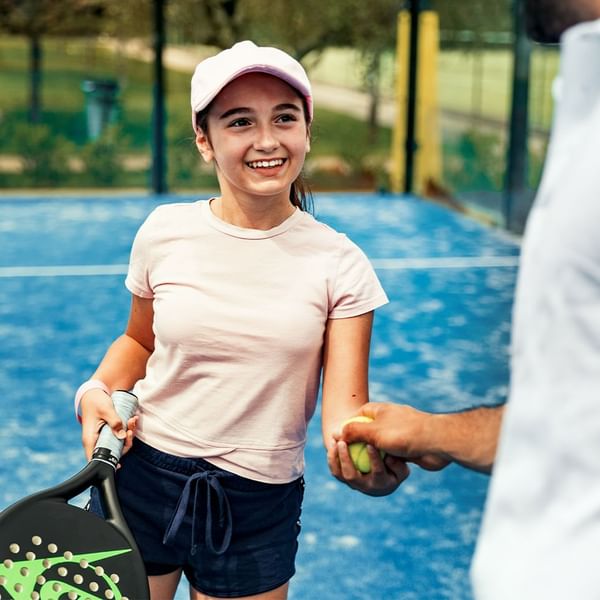 Ragazza con racchetta da tennis sorridente mentre un uomo le dà una pallina da tennis.