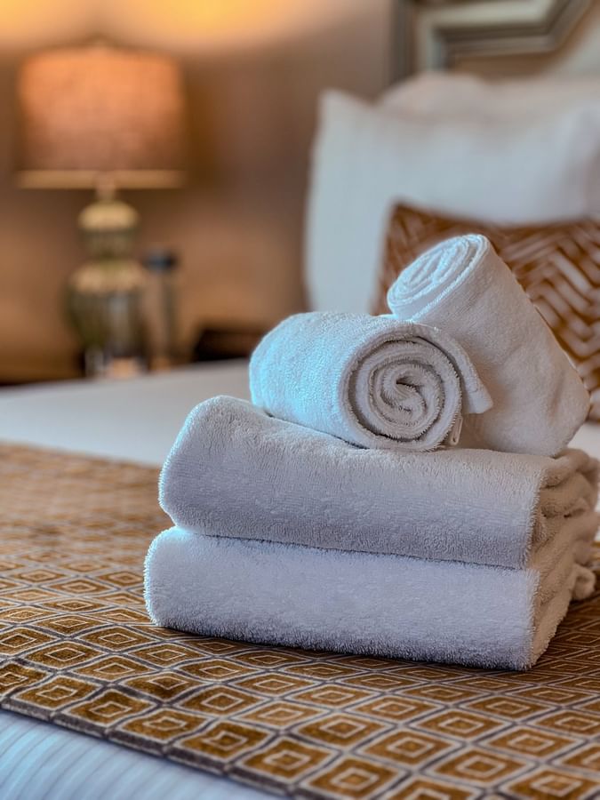 Rolled white towels on a bed with a patterned runner by a soft pillow in Deluxe Suite Accessible at Warwick Melrose Dallas