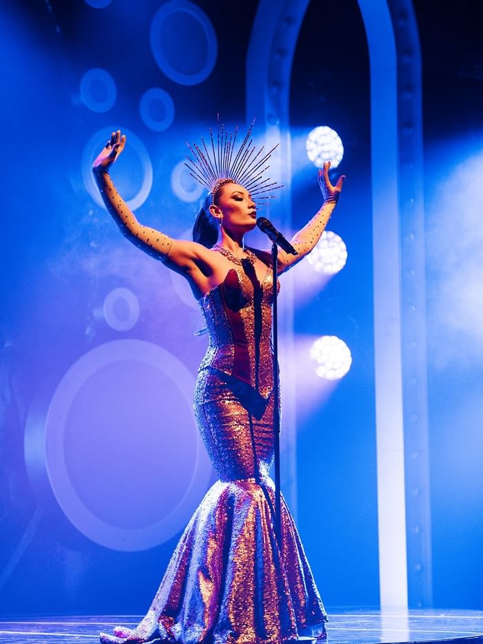 Performer in sequined mermaid gown and sunburst headpiece on stage with blue lighting for Blanc de Blanc Encore.