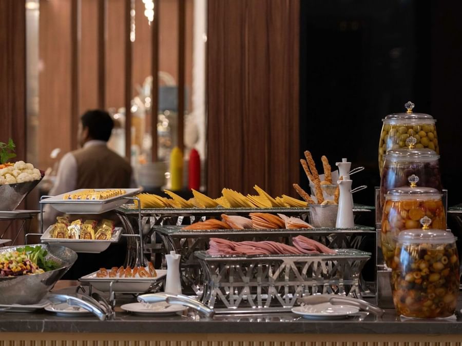 Cold cuts and cheese slices on silver stands by jars of olives at a breakfast buffet at Warwick Al Jubail