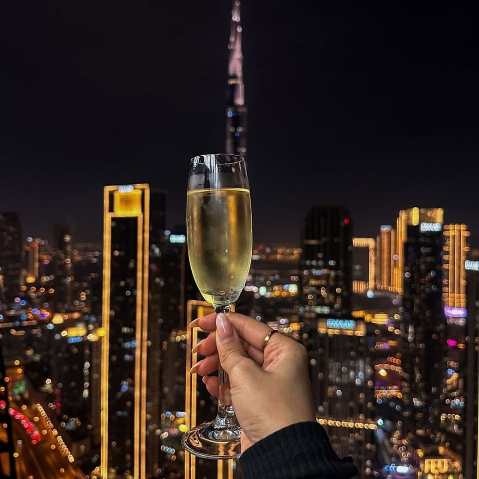 Hand holding a champagne flute against the glowing Dubai skyline at Malibu Sky Lounge & Bar with Burj Khalifa dining options
