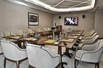 Grey chairs by a wooden table surrounding a large TV screen under bright lights in Kugu Hall at Warwick Ankara