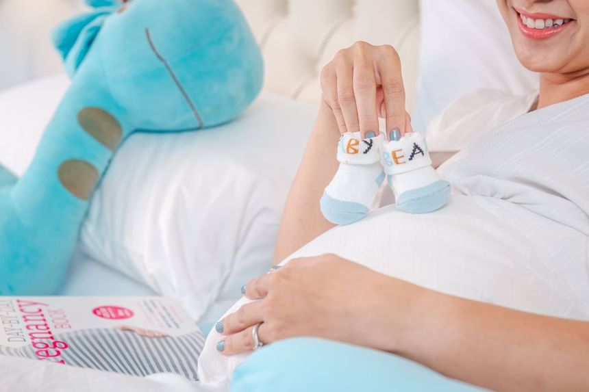 Pregnant woman holding baby socks while resting on a bed with a plush toy at Park Hyatt Saigon