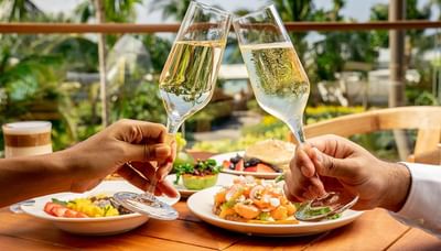 Two hands clinking champagne glasses in an outdoor setting, with gourmet dishes in The Residences at Seafire