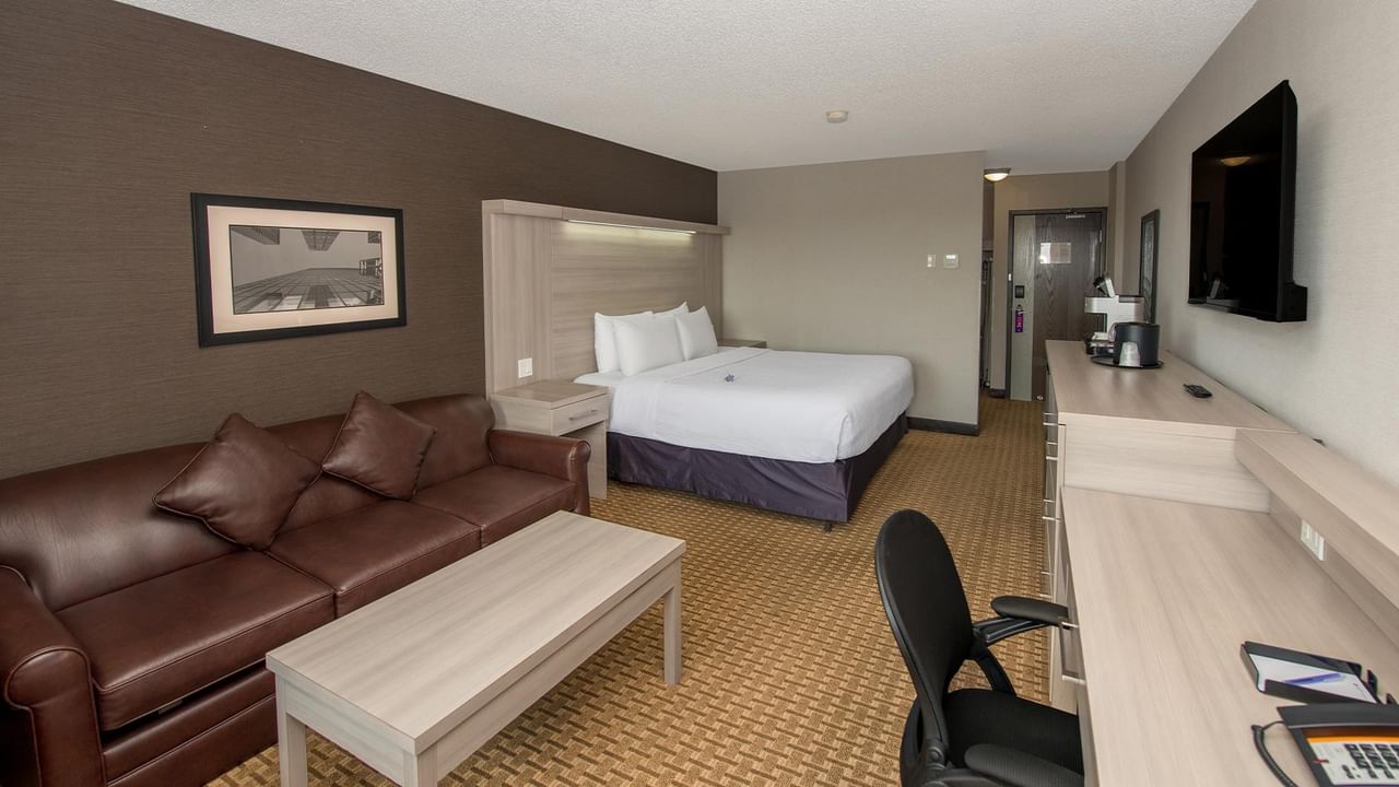 Spacious hotel room with white bed, brown leather sofa, work desk, and wall-mounted TV at Coast Calgary University Area Hotel by APA