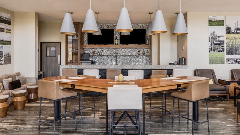 Modern bar area with a large wooden table, seating and stylish pendant lights at Fiesta Inn Suites Aeropuerto del Bajío