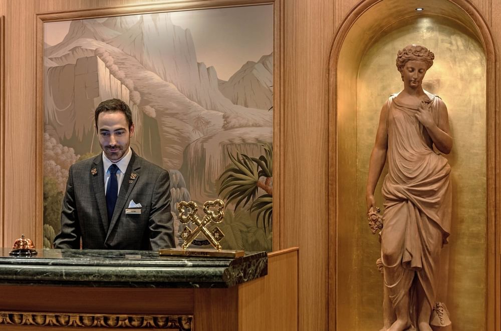 Concierge standing by a marble desk and a golden statue in a niche at the lobby of Hotel Westminster Paris