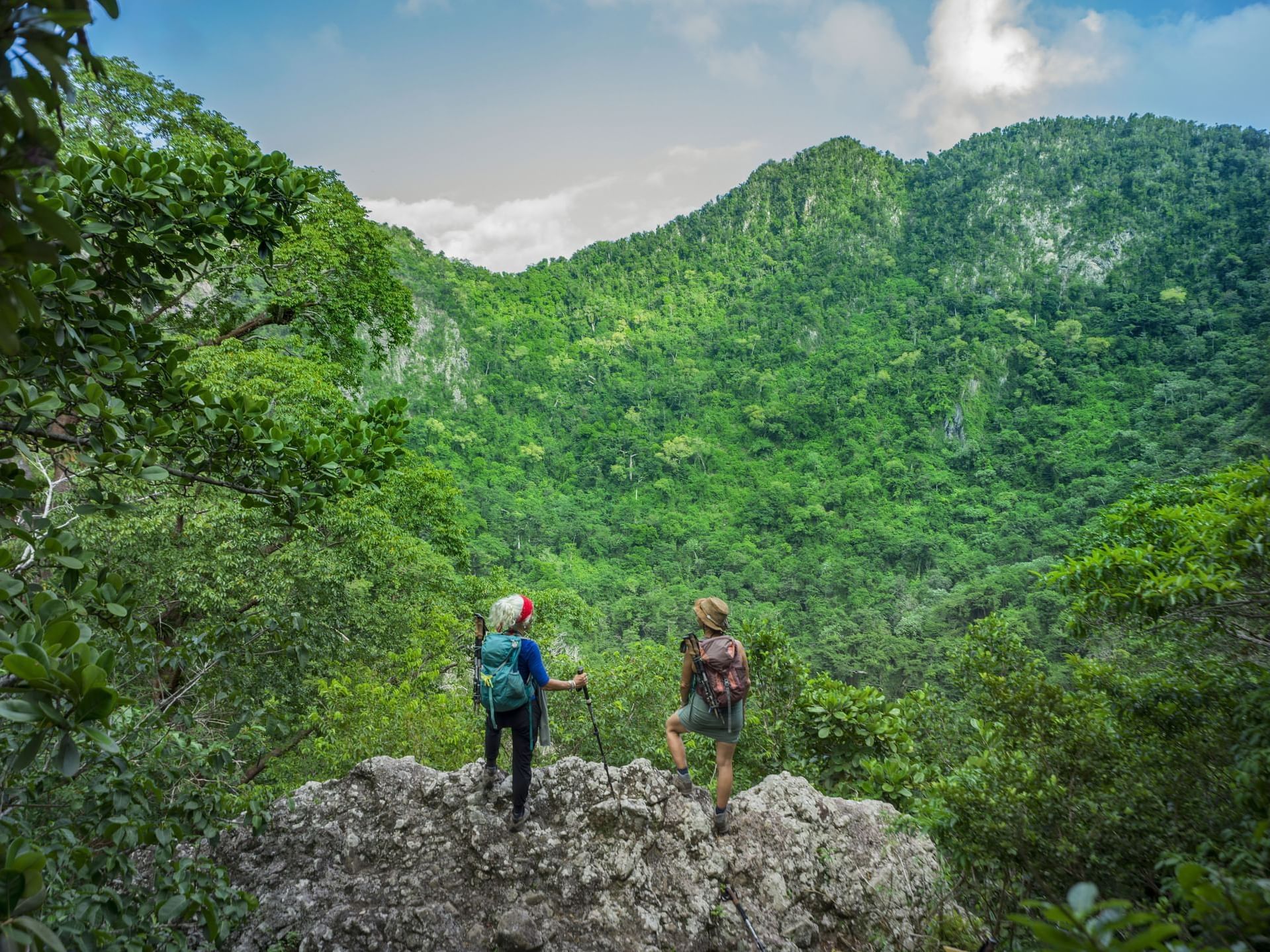 Two hikers stand on a rocky outcrop, gazing over a lush green valley surrounded by mountains near Golden Rock Resort