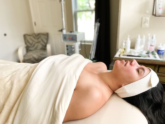 Woman lying on a bed with a towel covering her, wearing a white headband, in a spa setting.