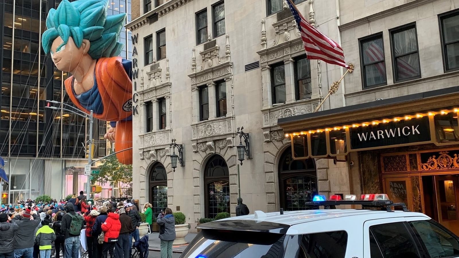 Large blue-haired Goku parade balloon watches a large crowd with police jeep in front of Warwick Hotels and Resorts