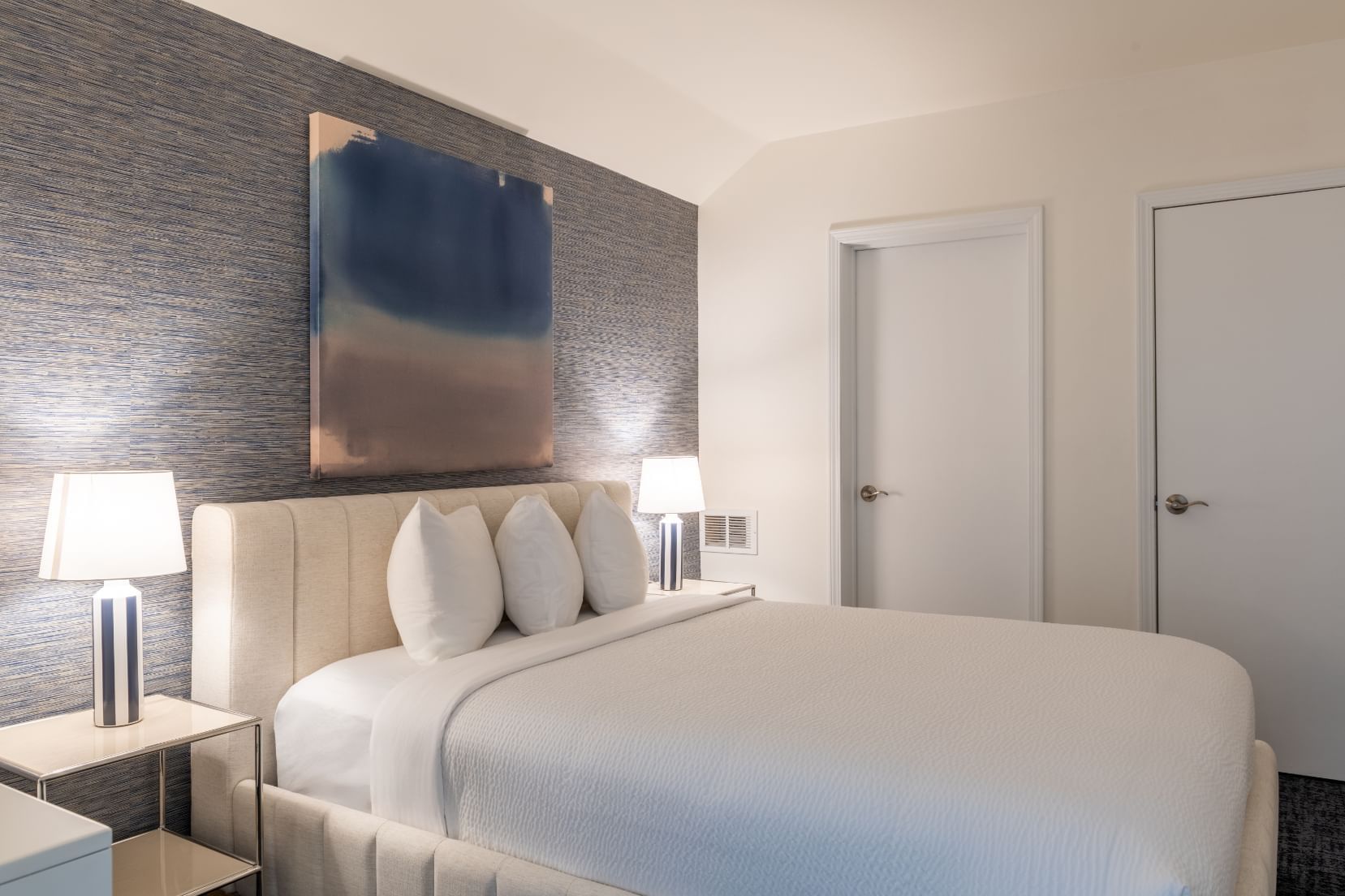 A neatly made bed with white linens and pillows, flanked by two nightstands with lamps, against a wall with abstract artwork.