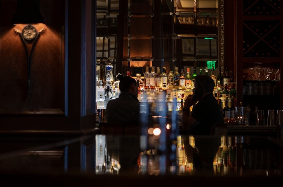 Guests at a bar with bottles under dim lighting surrounding The Library Bar at Warwick Melrose Dallas