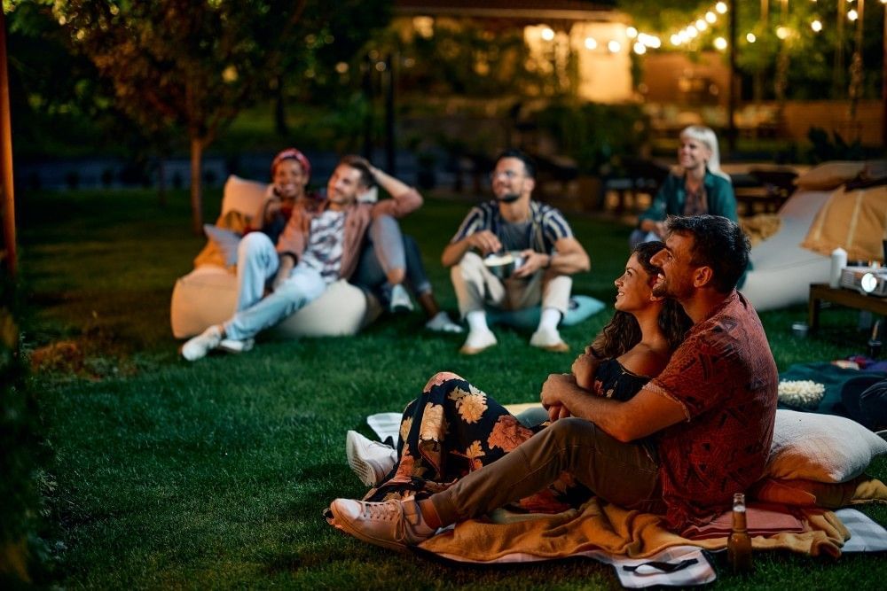 Couples sitting on blankets and beanbag chairs on an outdoor lawn with bistro lights overhead.