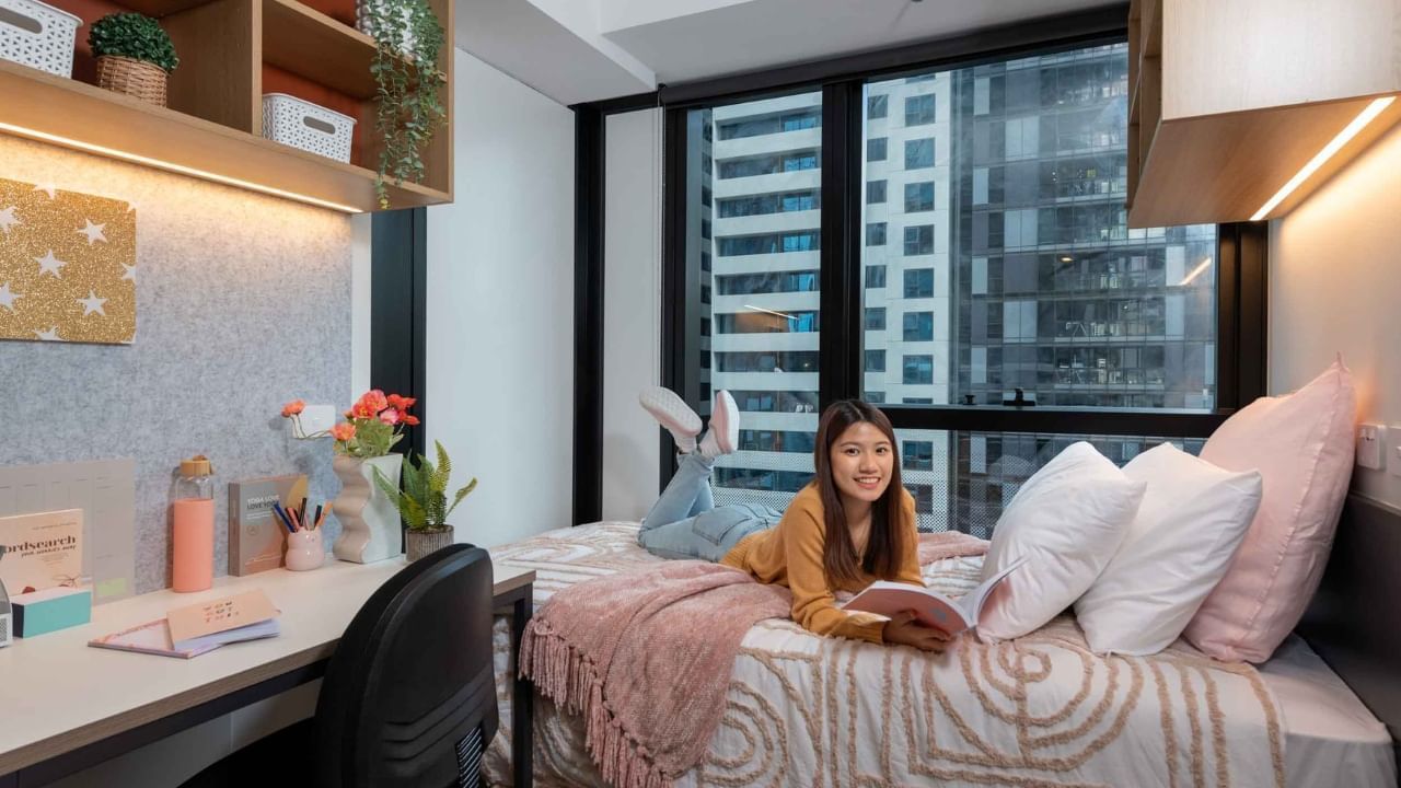 Resident reading book while sitting on bed with pillows and desk in studio room.