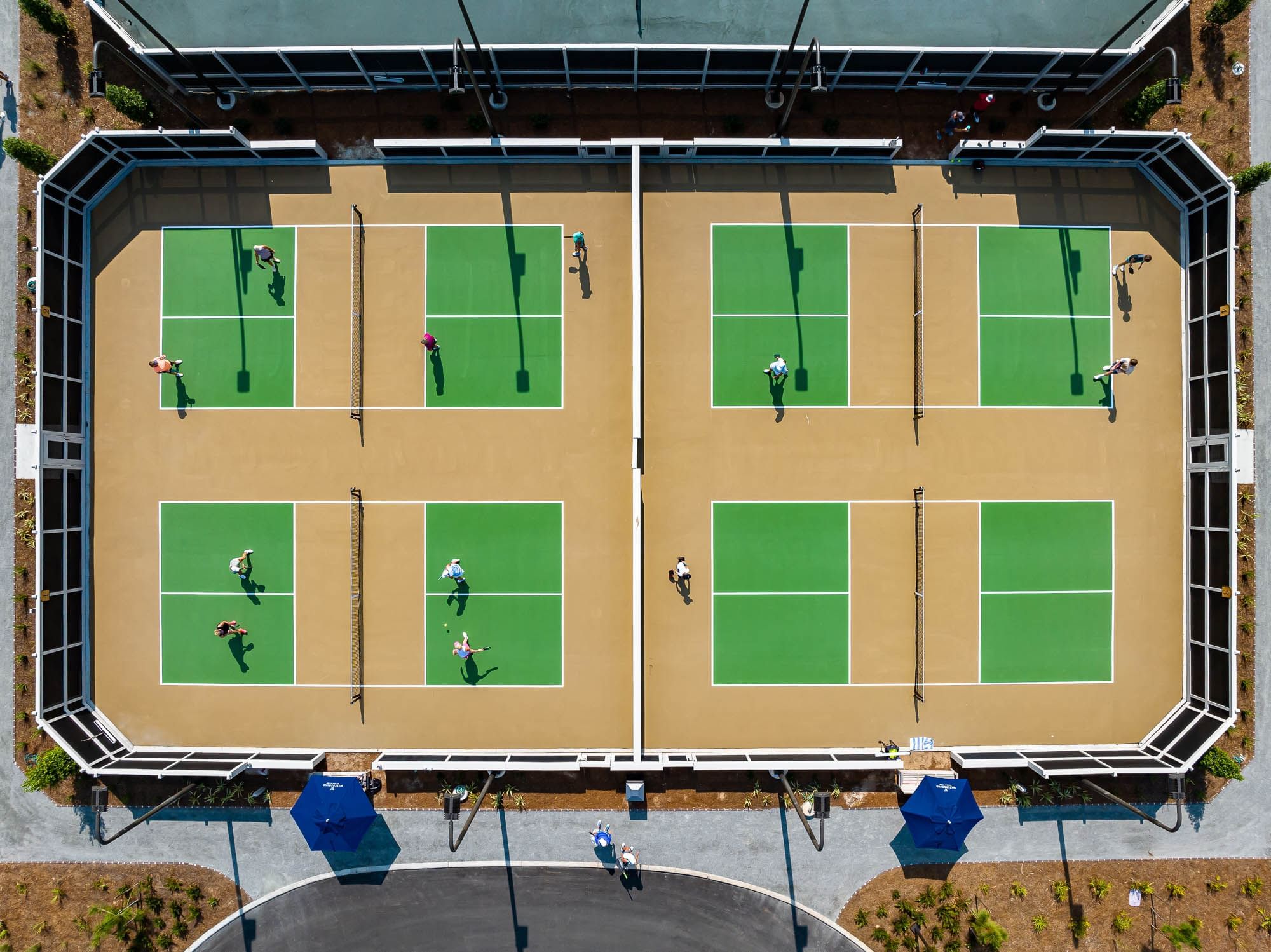 An aerial view of a tennis court with several people playing tennis and two blue umbrellas nearby.