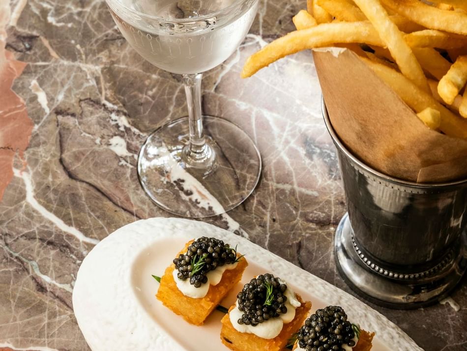 Stylish table setting at Granduca Houston with caviar appetizers, fries in a paper cone, and a cocktail on a marble surface