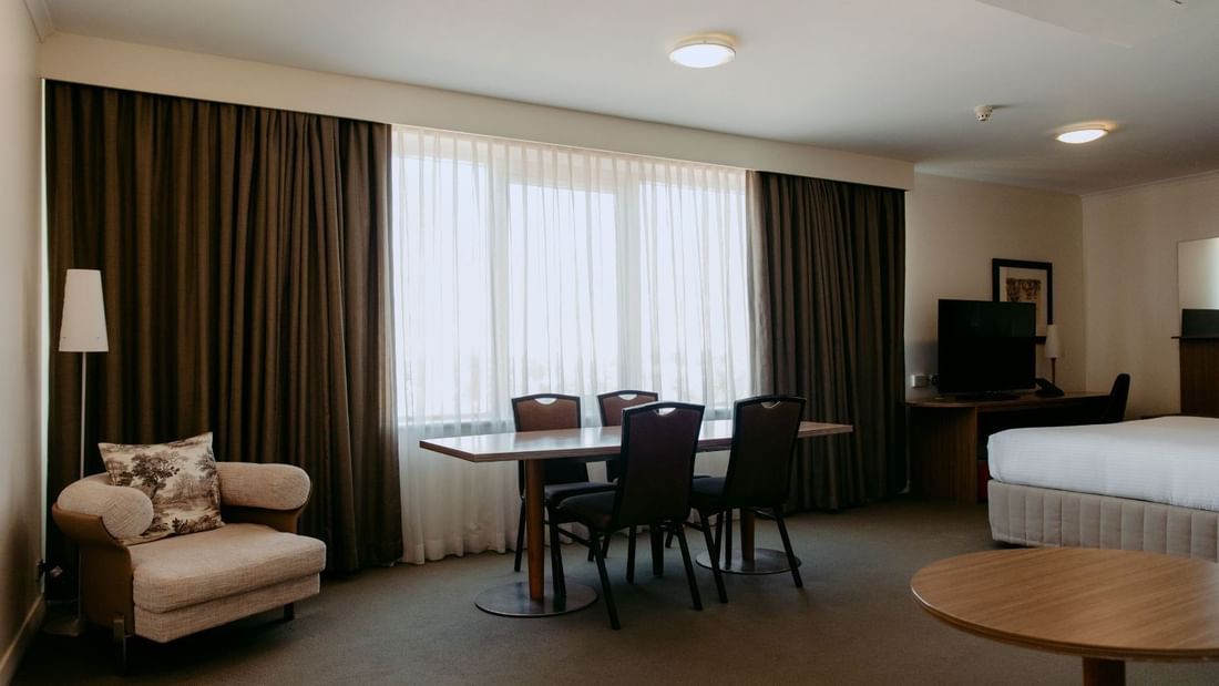 Hotel room with bed, desk, armchair, table with four chairs, and large window with curtains.