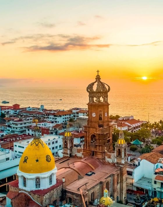 Ornate church tower and golden domes facing a city skyline and ocean during a glowing sunset at Los Arcos Suites