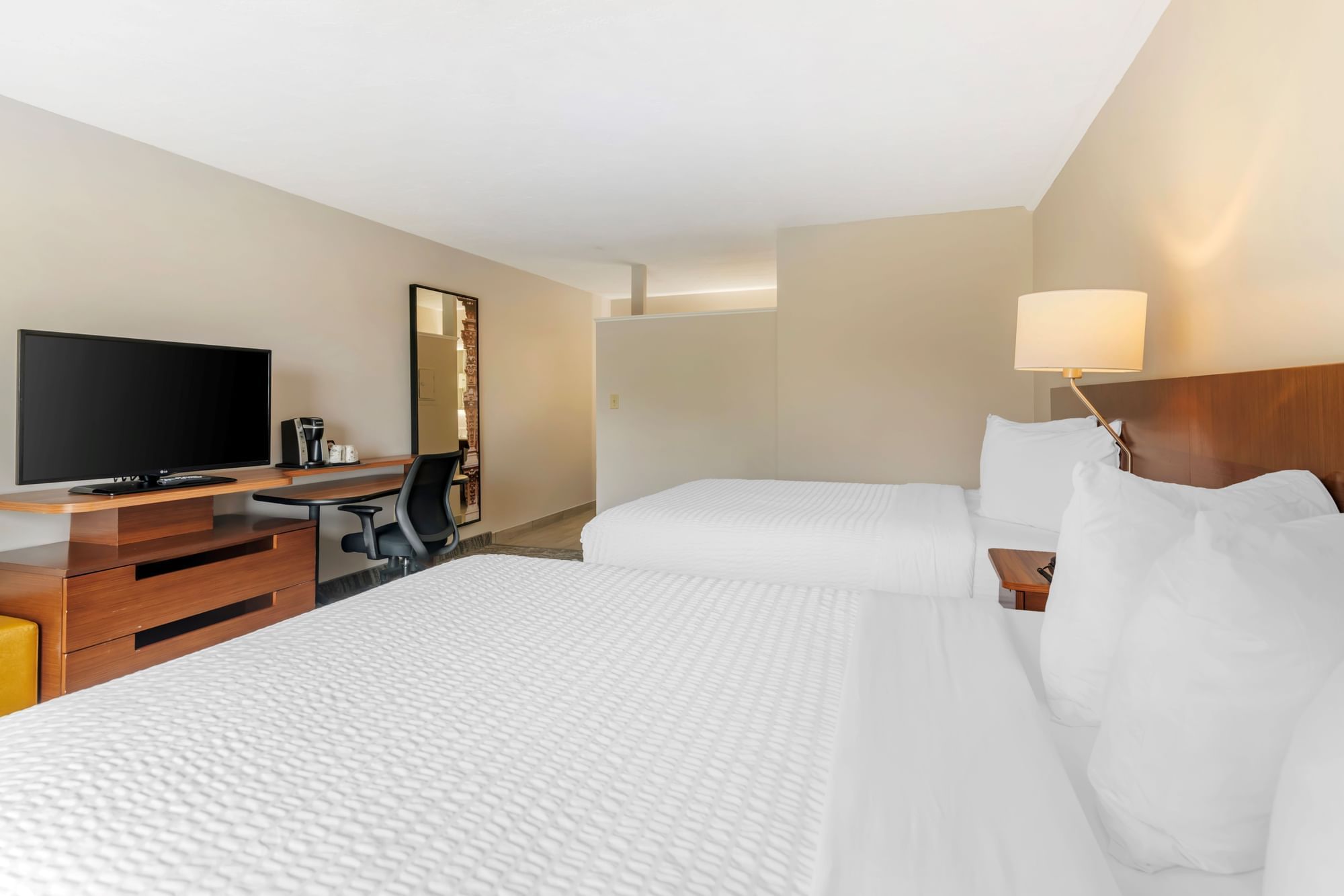A hotel room with two queen beds, a flat screen TV on a wooden dresser, a desk with a chair, and a lamp on a nightstand.