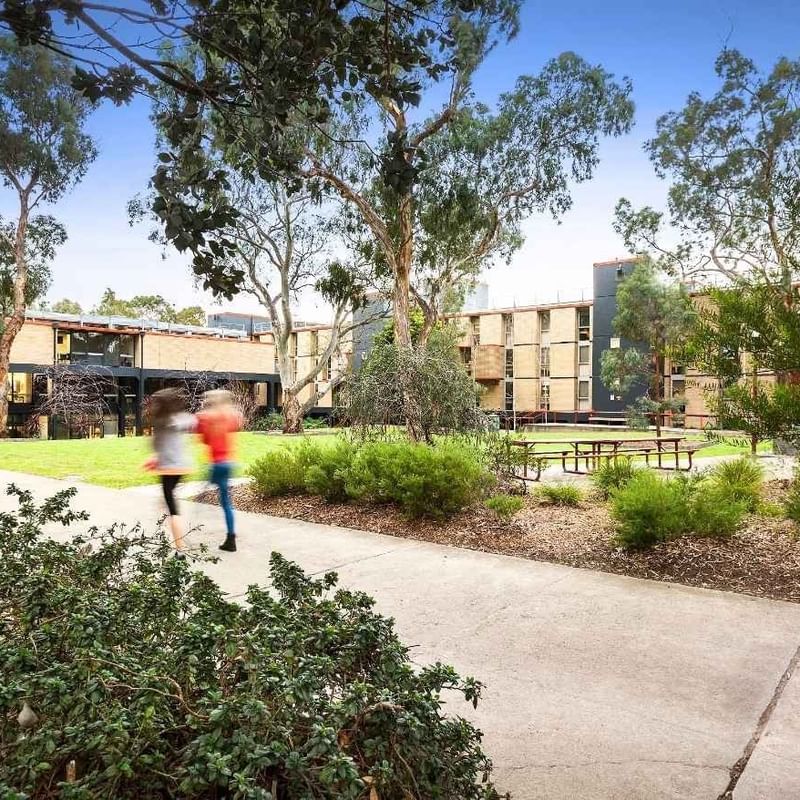 Two students running on a concrete walkway surrounded by trees and buildings at La Trobe University – Menzies College.
