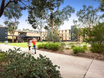 Two students running on a concrete walkway surrounded by trees and buildings at La Trobe University – Menzies College.