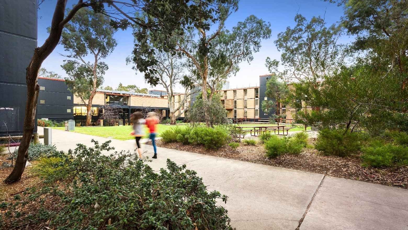 Two students running on a concrete walkway surrounded by trees and buildings at La Trobe University – Menzies College.