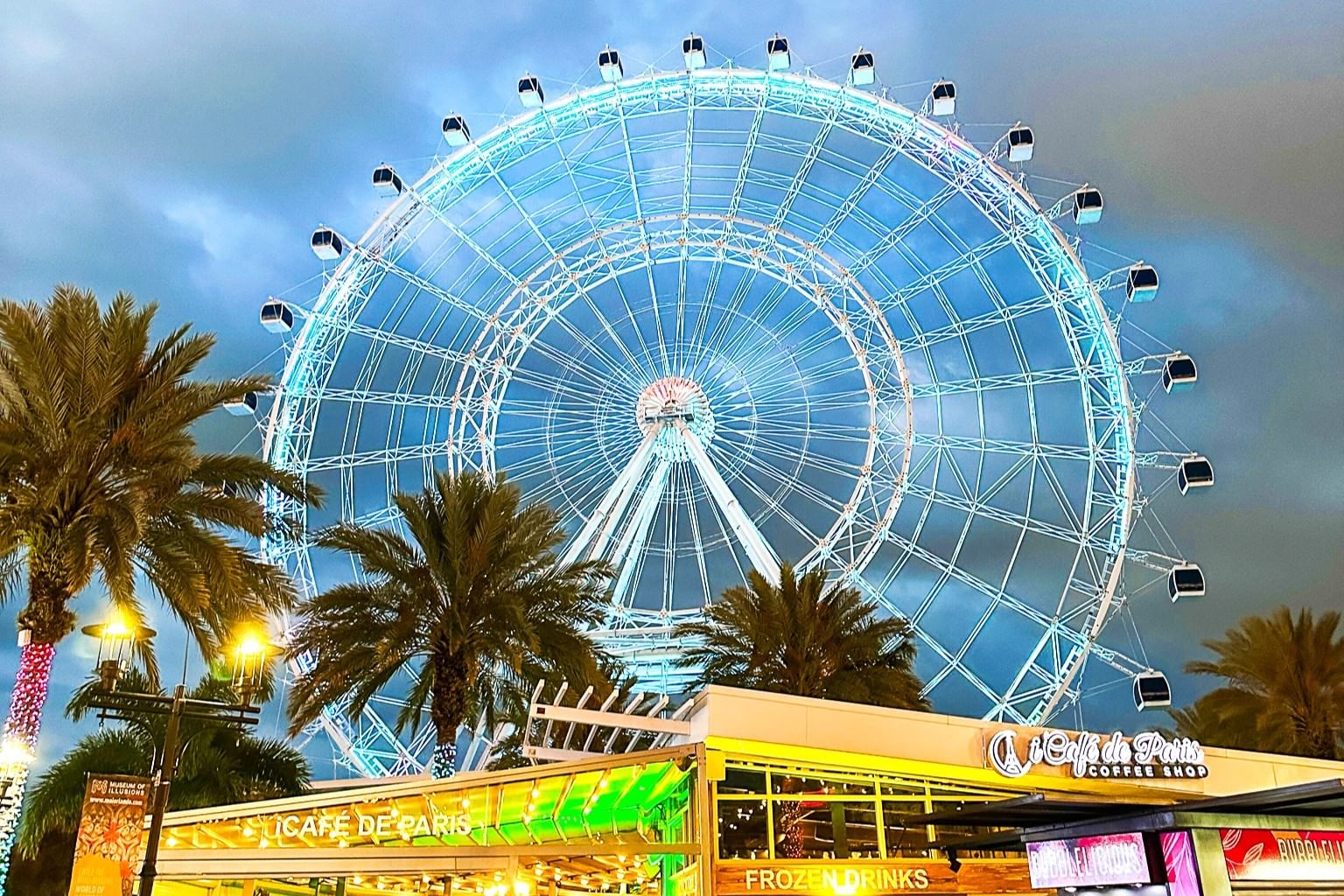 A glowing white ferris wheel lit with colorful LED lights against a cloudy twilight sky with glowing buildings and palm trees below.