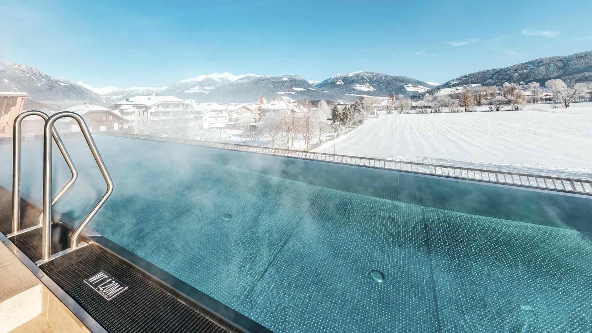 Outdoor infinity pool with steam overlooking snowy mountain landscape at Falkensteiner Hotel Kronplatz