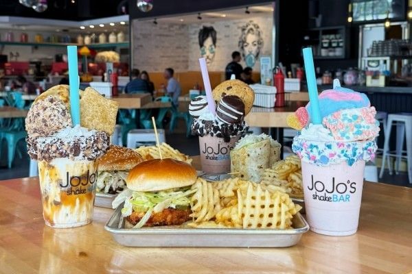 Three colorful and enormous milkshakes sit on a table beside trays with burgers and waffle fries at JoJo's ShakeBAR in Orlando.