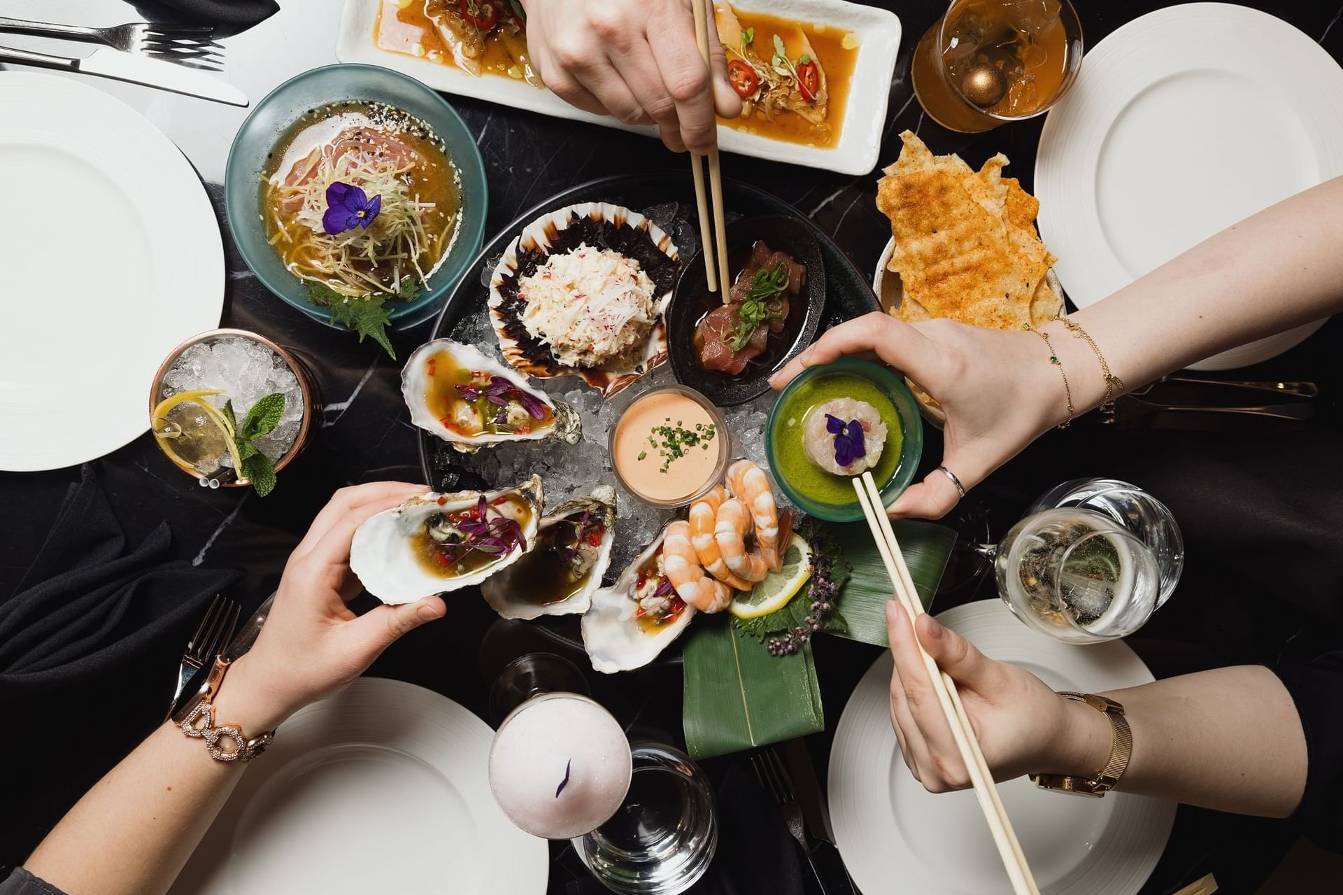 Top-down view of a seafood platter with oysters, shrimp, and sushi by drinks and table settings at The Kitchens