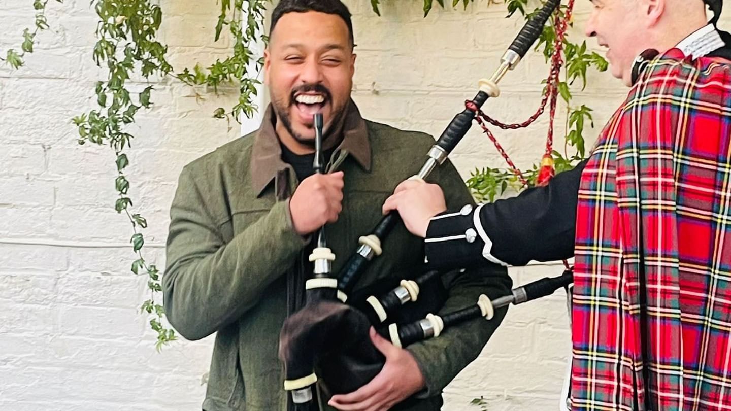 A person in a tartan kilt plays the bagpipes beside ivy-covered walls near Seaton House