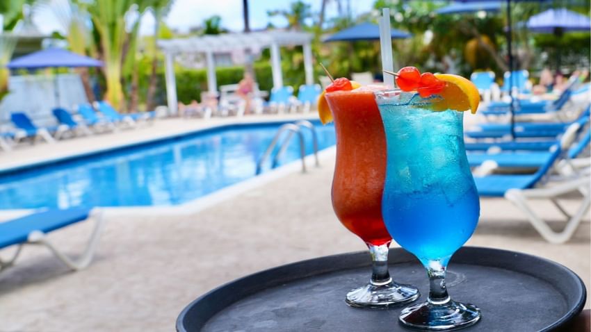 Two colorful tropical drinks on a tray by a clear swimming pool under bright skies, surrounded by seats at Time Out Hotel