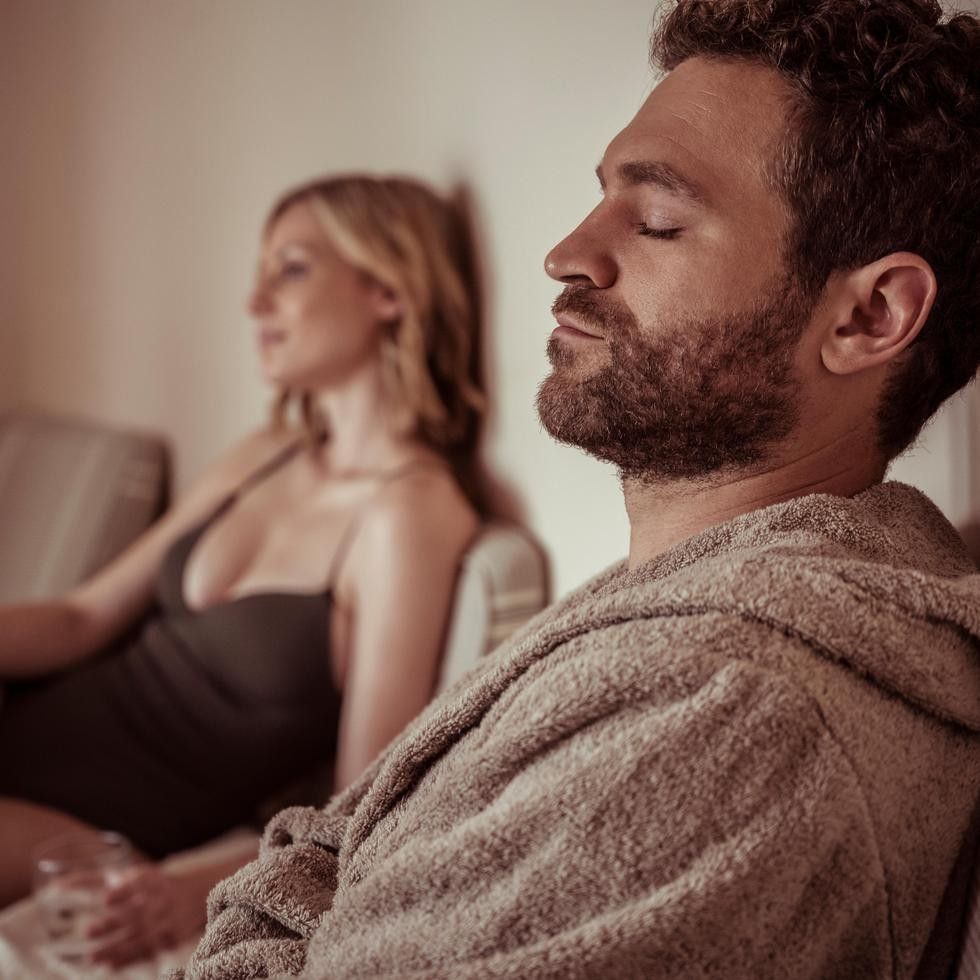 Man in bathrobe relaxing with eyes closed while woman sits in background during spa experience
